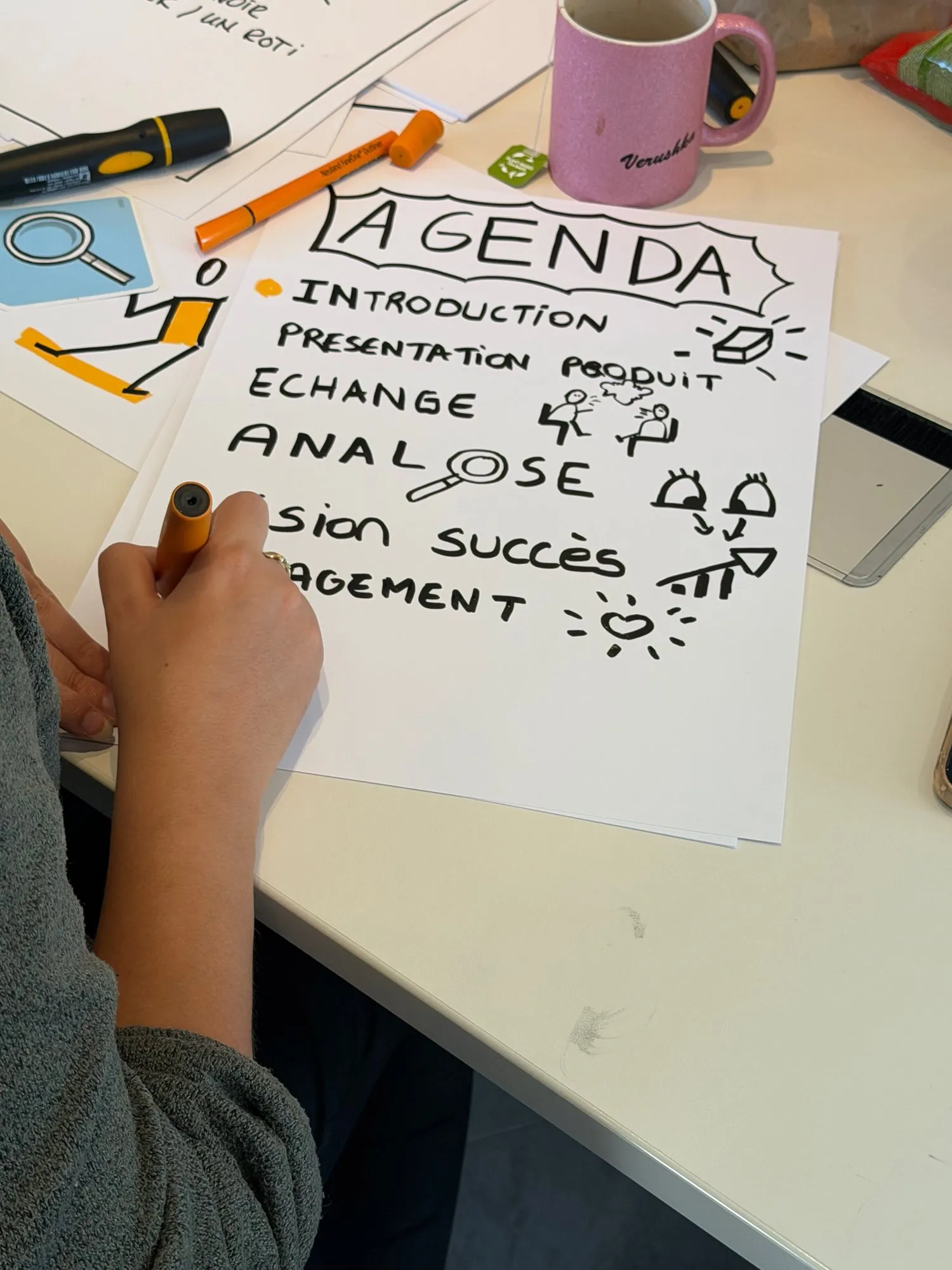 Une main tient un feutre orange au-dessus d’une feuille titrée « Agenda », où chaque ligne a son pictogramme