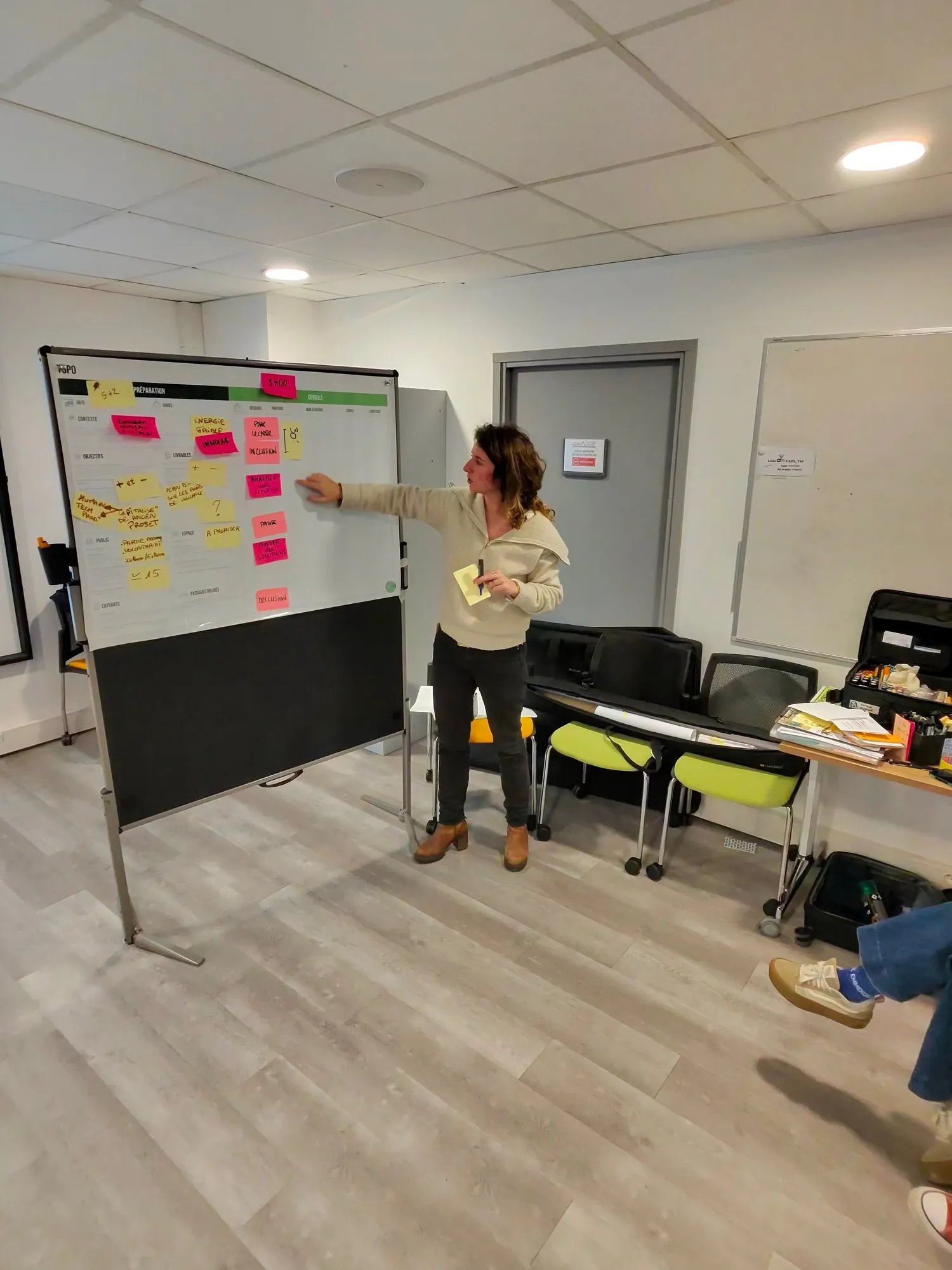 Une participante debout montre du doigt un panneau couvert de post-its, son conducteur d’atelier