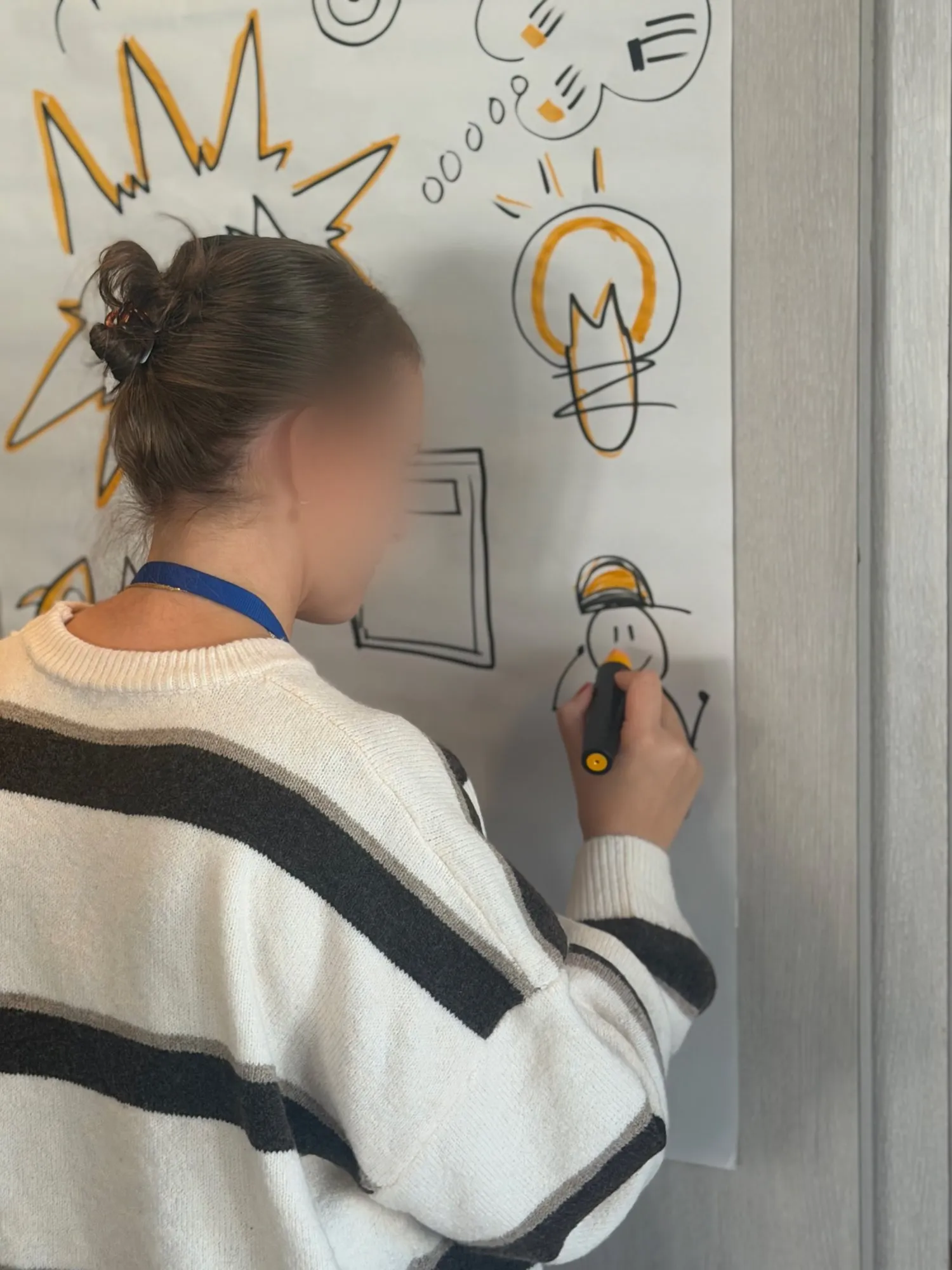 Une participante, de dos, trace un personnage au feutre sur un paperboard déjà couvert d’une ampoule et d’un éclat orange